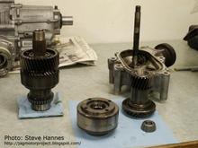 Steve Hannes: First the small shaft (Driven). It is now on the right in this picture. It is a solid shaft containing two gears, the upper gear gets its drive from the main shaft, transferred to the bevel gear just visible at the base of the shaft. Again, this is the torque delivery (the 60%) to the rear wheels. Now for the larger shaft assembly (on the left in this pic). This assembly has a lot going on. In this picture I've removed the upper assembly. It now is positioned in the forefront and turned upside down. It is the sealed viscous coupler unit. It is driven on the mainshaft by four dowel pins and resides right above the mating primary drive gear for the rear wheels.
***** more info: http://jagmotorproject.blogspot.com/2007/02/motor-project-17-transfer-time.html   *****