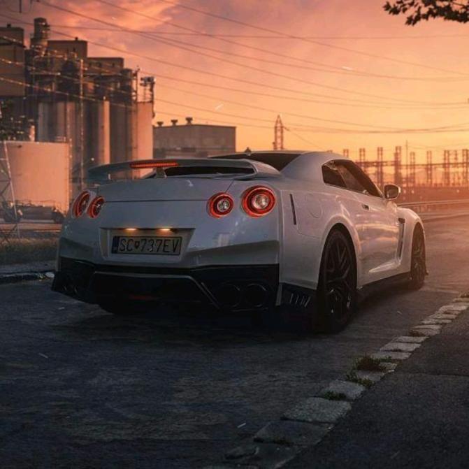 A white sports car is parked on a road during a sunset with an industrial background.